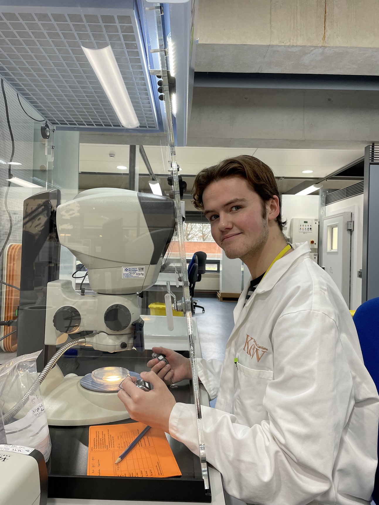 Rowan wearing a white lab coat smiling at the camera. He is sat infront of a microscope in a dusthood, which has a germination plate under it.